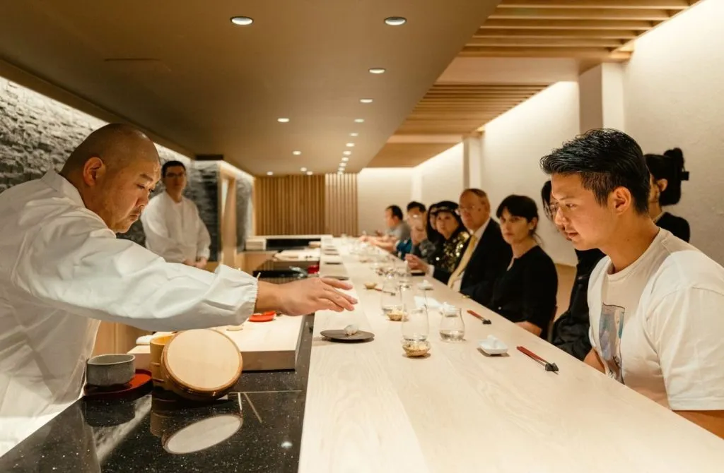 A skilled sushi chef meticulously preparing a delicate nigiri piece at an intimate wooden omakase dining counter.